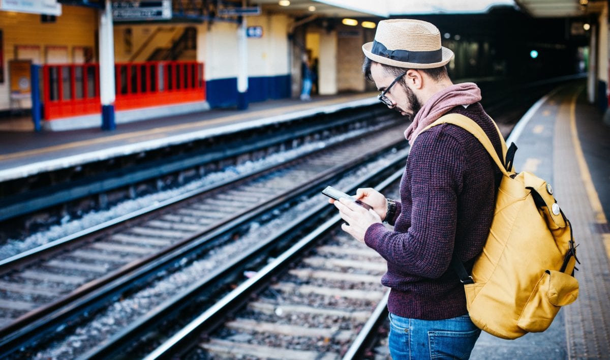 A man with a backpack and hat waiting on a train station platform.