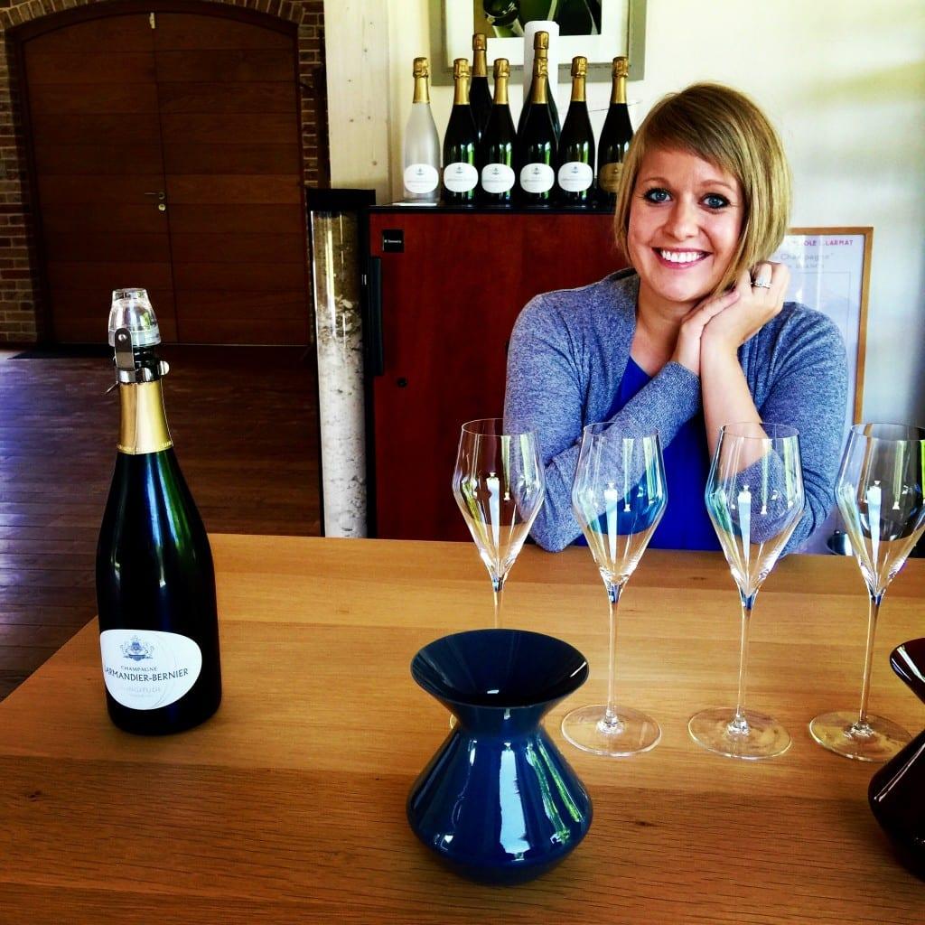 A woman sitting at a table with wine glasses and smiling at the camera, with Champagne