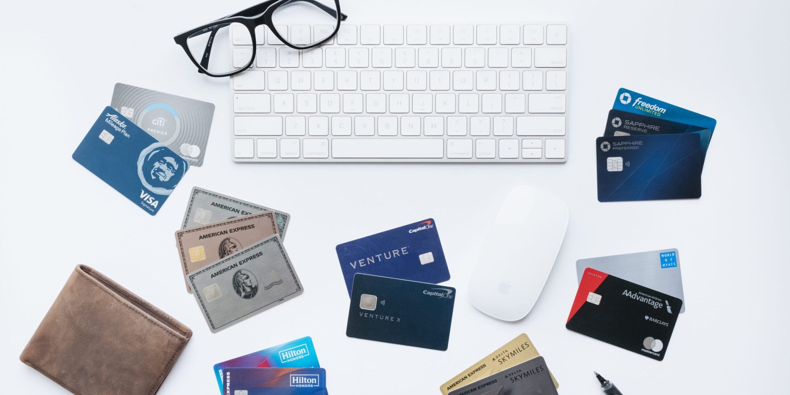 Credit cards spread out on a desk with a pair of glasses, a wallet, keyboard, and mouse.