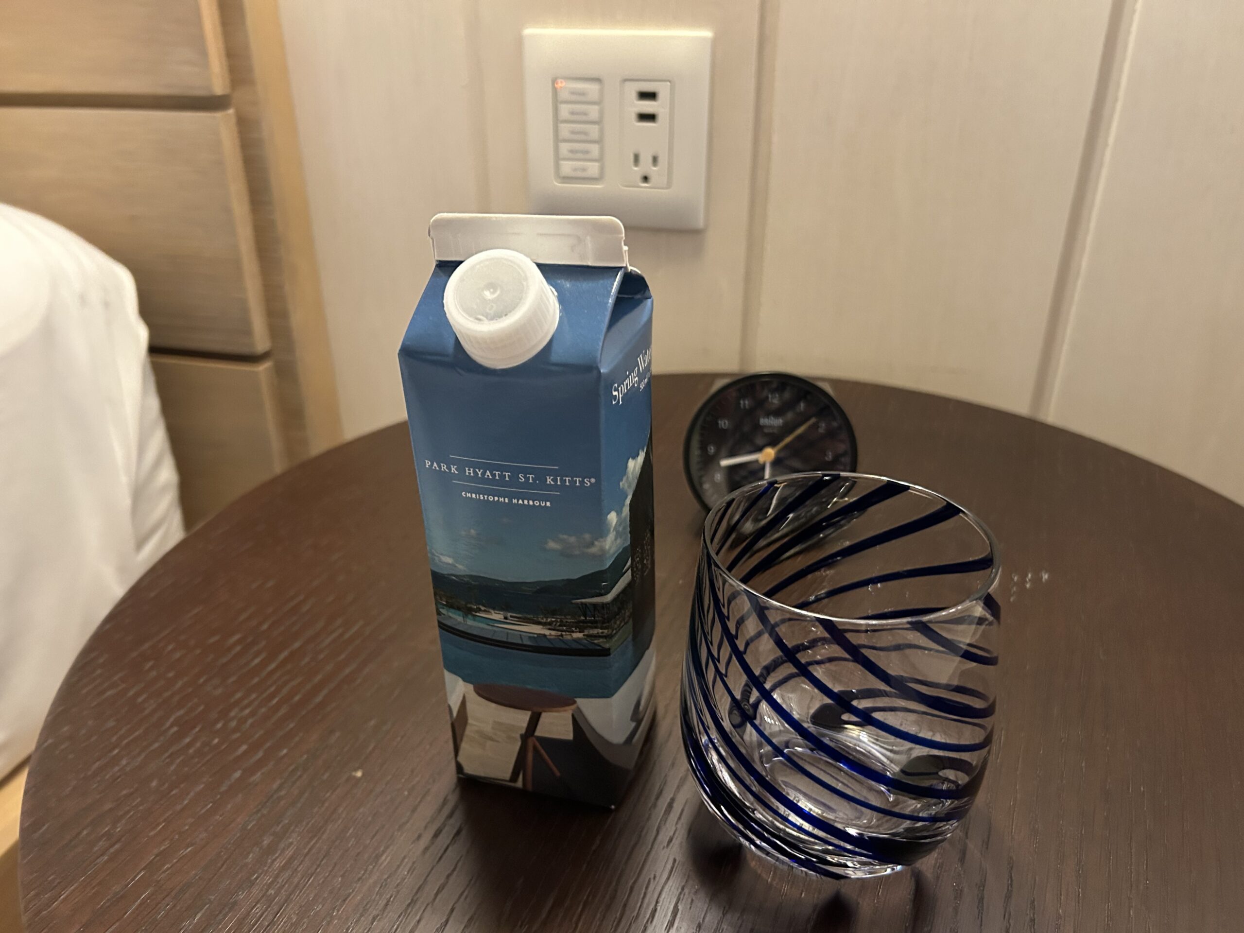 Park Hyatt St. Kitts-branded cardboard water bottle with a striped water glass on a bedside table