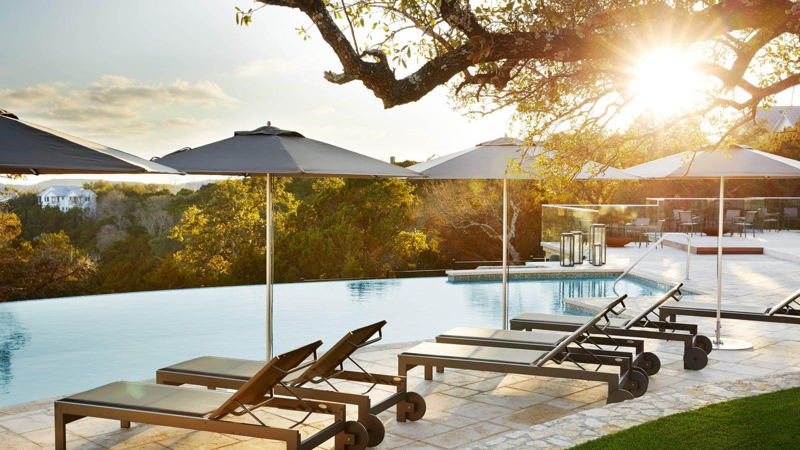 Lounge chairs with umbrellas sitting by an infinity pool with the sun shining through the trees. 