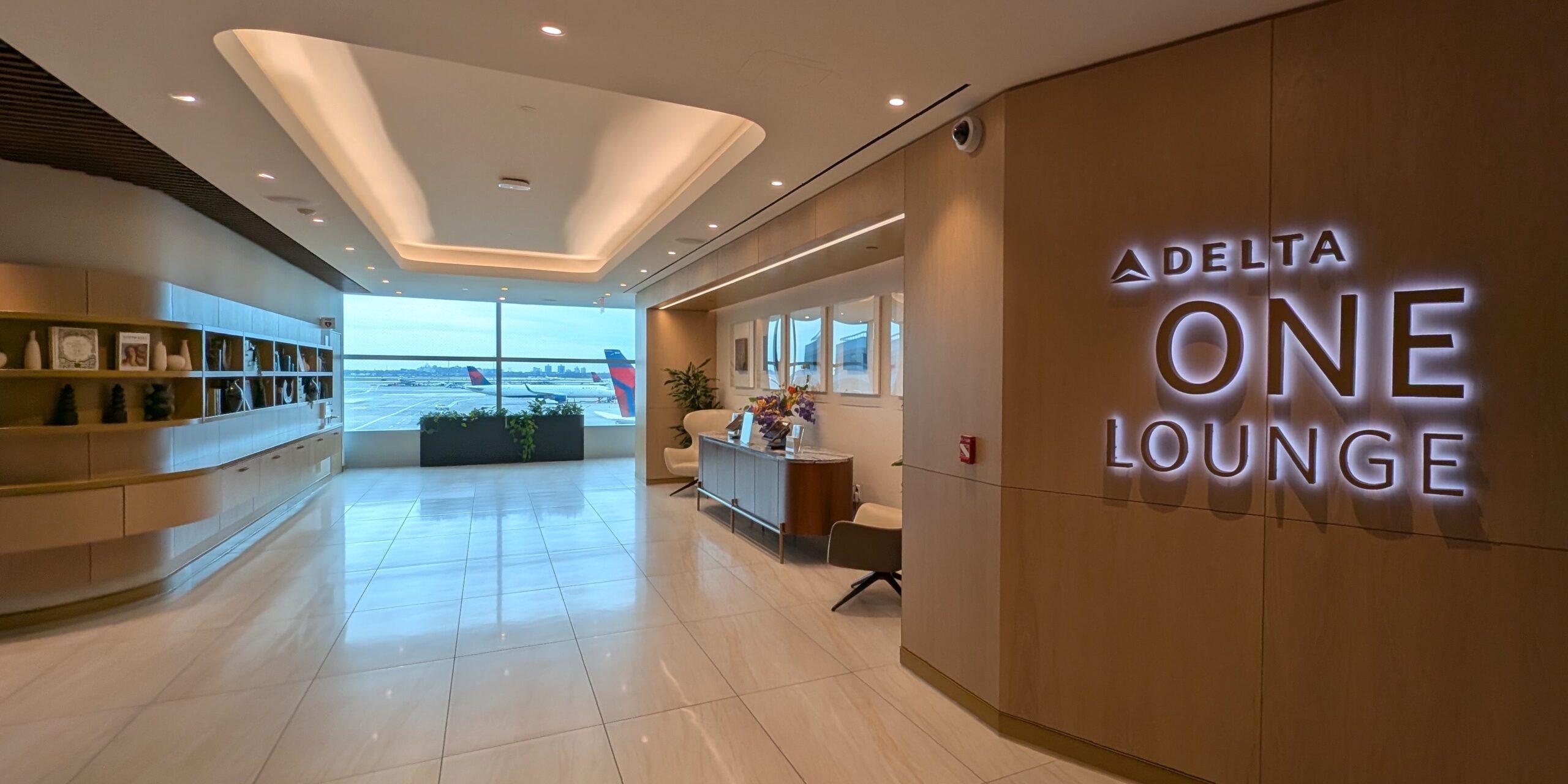 Entrance to the Delta One Lounge at JFK with a bookshelf on the left side and Delta One signage on the right. Two Delta airplanes sitting on the tarmac through the window at the end of the entrance. 
