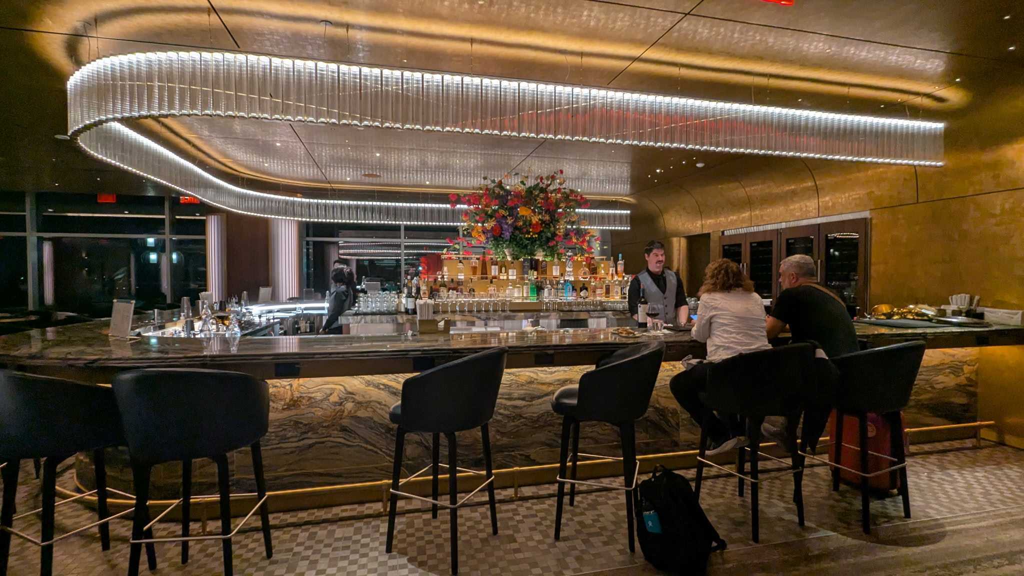 Two patrons seated at the gold-trimmed horseshoe-shaped bar with marble finishes and a lighted chandelier at the Delta One Lounge JFK. 