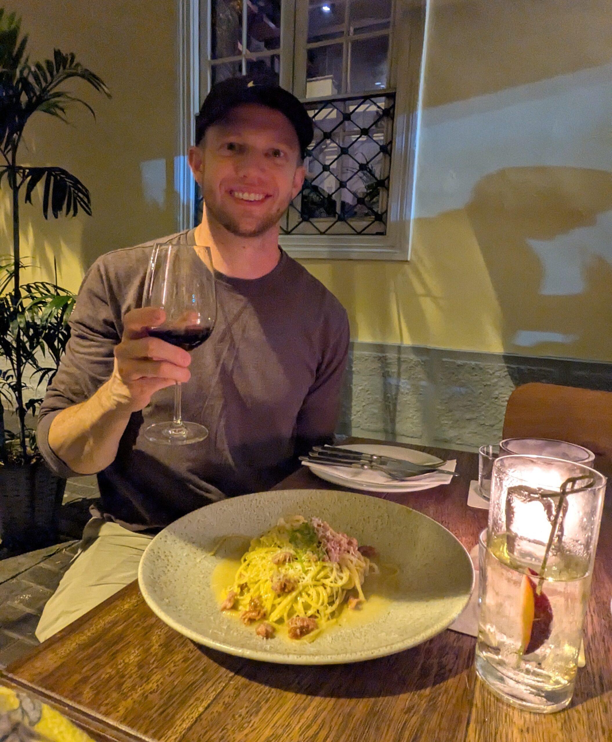 Thrifty Traveler's Jackson Newman raising a glass or red wine with a plate of pasta on the table in front of him. 