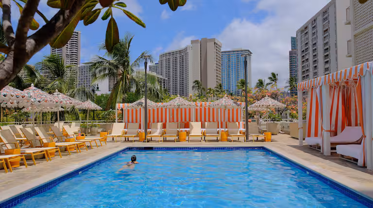 The Ambassador Hotel of Waikiki roofdeck pool with cabanas and lounge chairs, surrounded by palm trees and Honolulu's skyline. 
