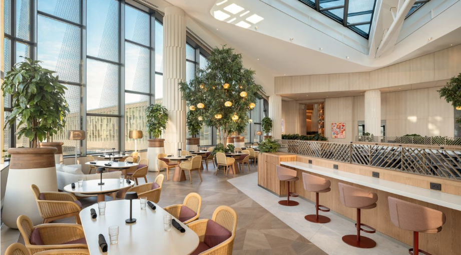 After a 3-Year Wait, Capital One’s 'Flagship' Landing Is Ready to Open at LGA 2 Capital One Landing (LGA) Dining Room with tables and chairs, fake plants, and countertop seating, soaked in natural light.