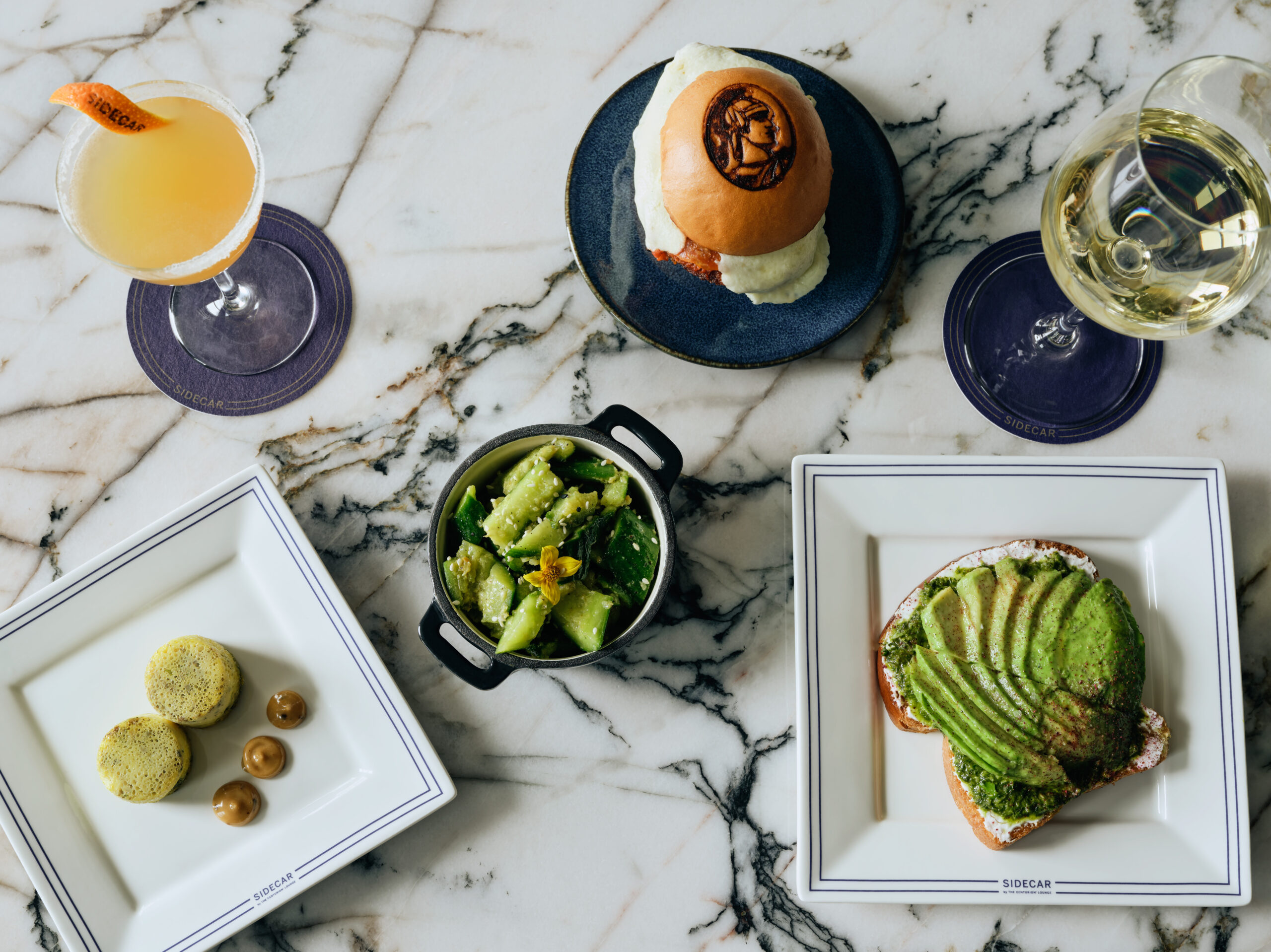 Amex Opens New ‘Sidecar' Lounge Concept at LAS ... & It’s Tiny 3 Small plates of food and two drinks on a marble countertop.