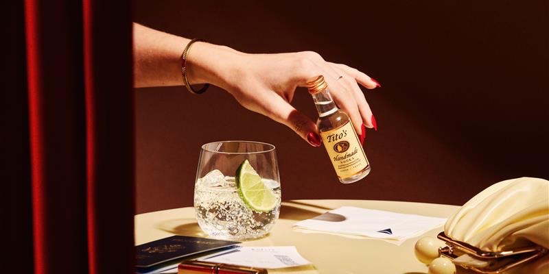 New: T-Mobile Customers Get a Free Drink on All Delta Flights 1 A hand holding a mini bottle of titos with a passport, coin pouch, and Delta napkin on a table