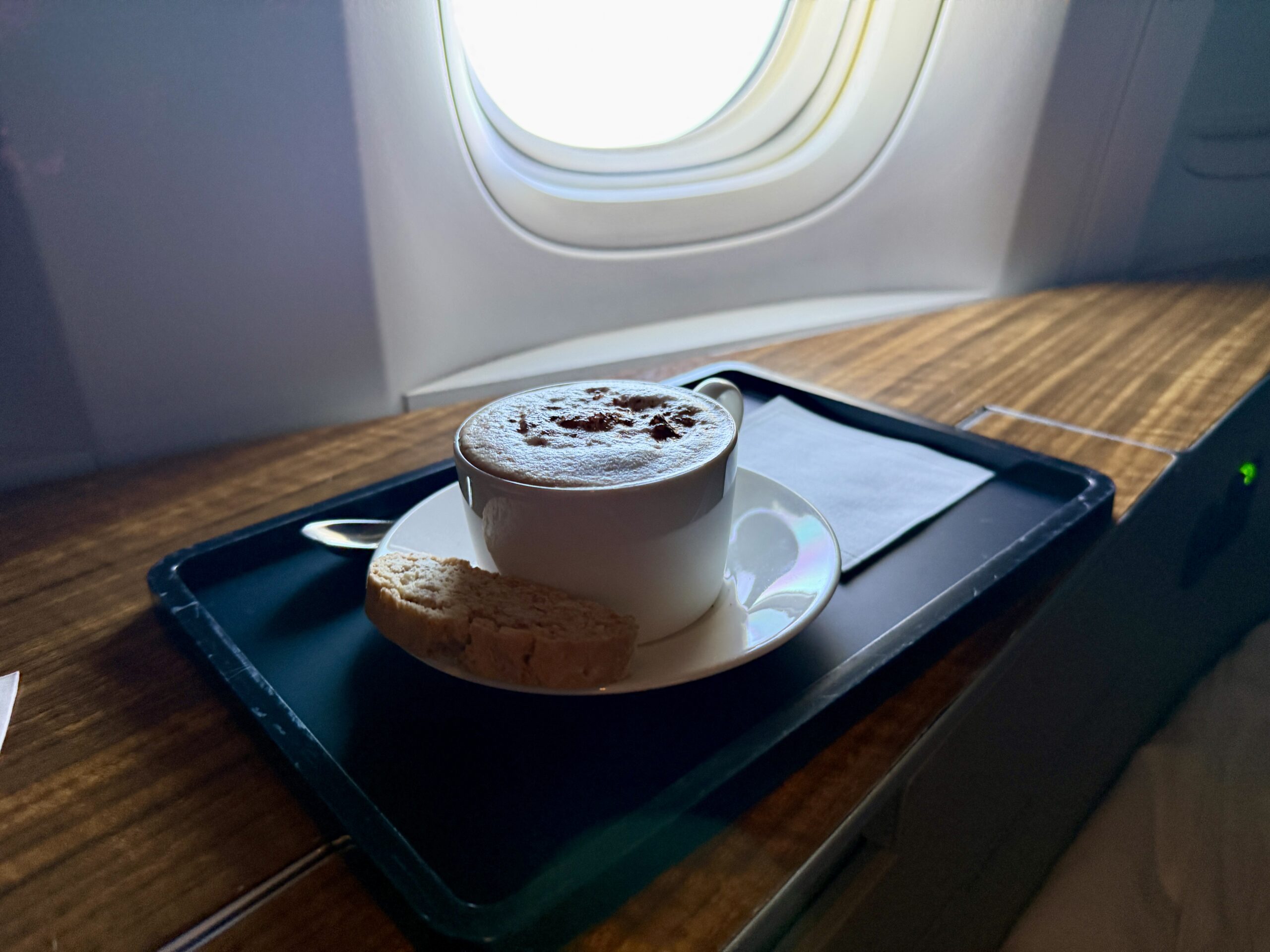 Aging Like a Fine Wine: Cathay Pacific First Class 777 Review 37 cappuccino with foam sitting on a small black tray with open plane window