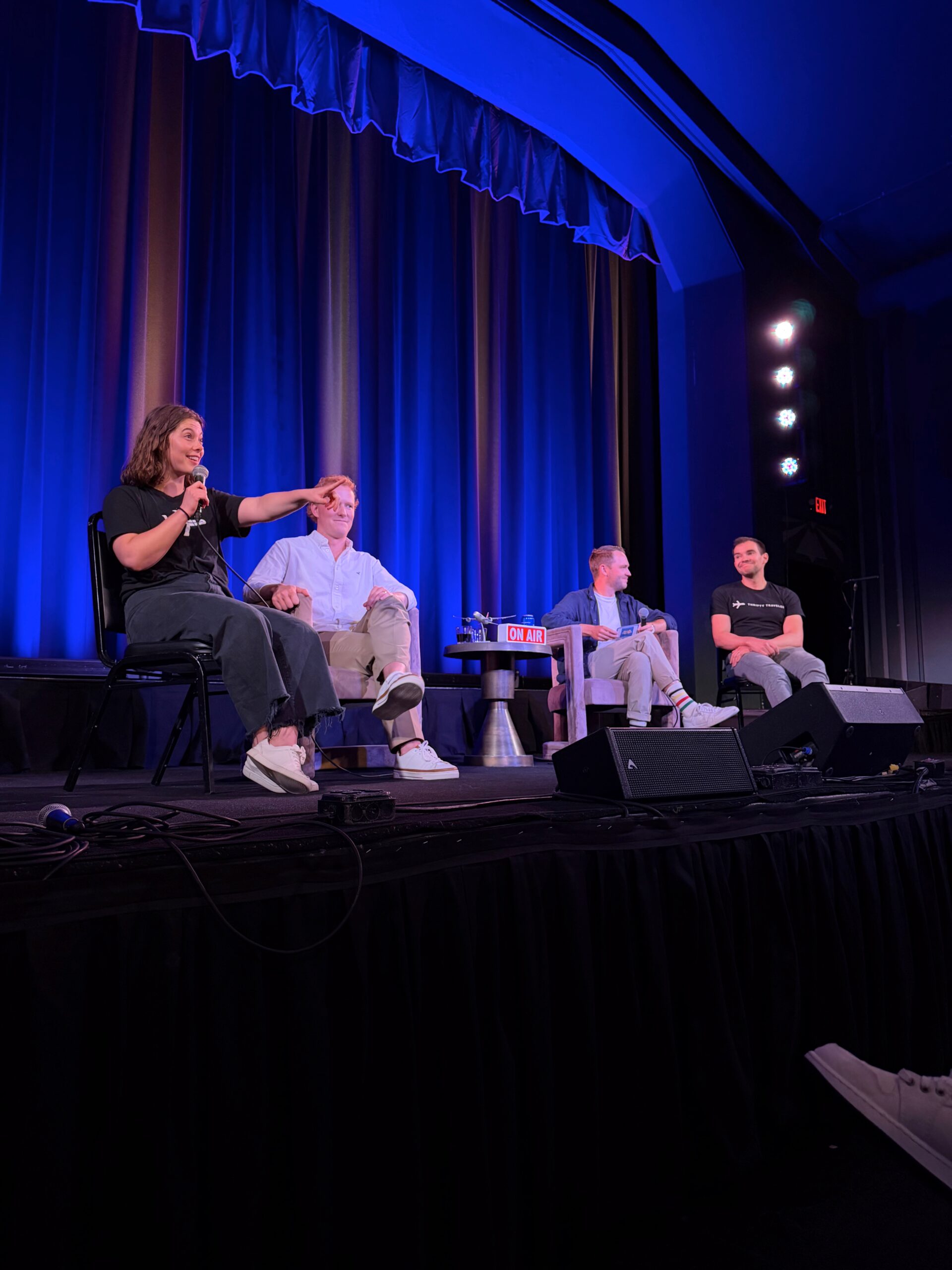 Recapping Our Live Podcast: Travelers Pack the Parkway for Hot Takes & Humiliation 4 woman on stage pointing with mic in hand alongside three men sitting